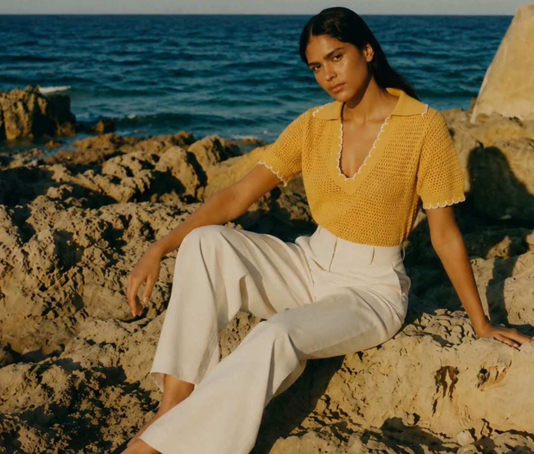 Woman wearing an An’ge yellow knit top with white trousers, sitting on coastal rocks by the sea in a chic and elegant summer fashion look.