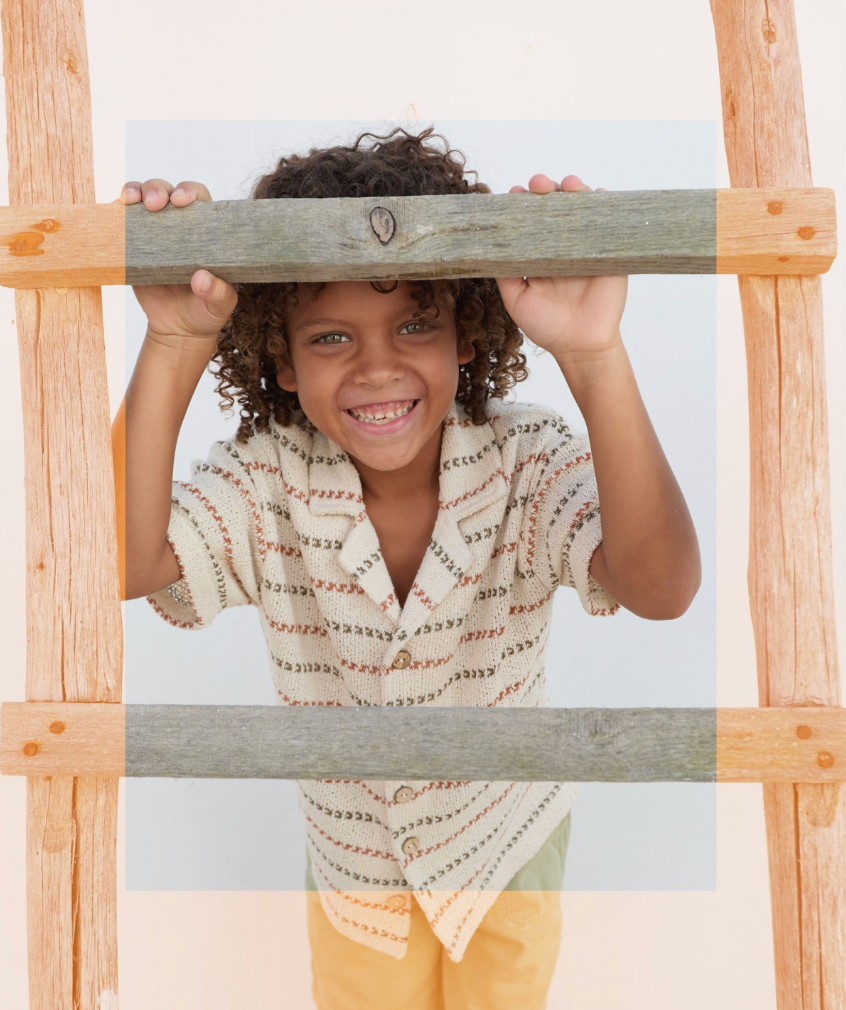Curly-haired child smiling through a wooden play frame wearing a soft knitted striped shirt and yellow shorts.